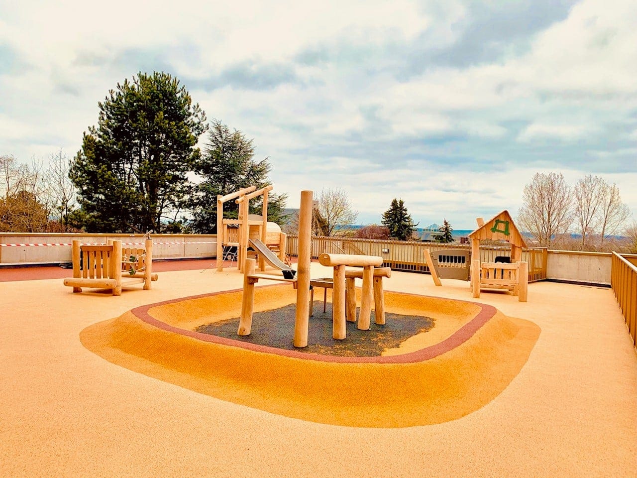 Aire de jeux en bois naturel avec toboggans et équipements ludiques sur un sol en revêtement souple, située en extérieur.