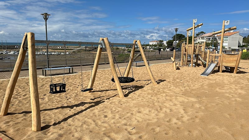 Aire de jeux en bois sur une plage, avec balançoires et structures de jeu pour enfants, en bord de mer.